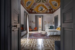 A room with a patterned floor, a bed with yellow pillows, a chair, a desk, and a ceiling with ornate designs. - Teatro Bellini, suite centro storico [Alcova L.] (Catania)