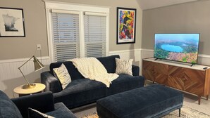 A living room with a dark blue sofa, a matching ottoman, a wooden TV stand, and a large flat-screen TV.