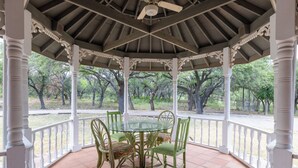 A gazebo with a glass table and chairs, surrounded by white railings and columns, overlooking a garden with trees. - Charlie IV House at Y.O. Ranch Headquarters - 4-Bedroom Ranch Stay (Mountain Home)
