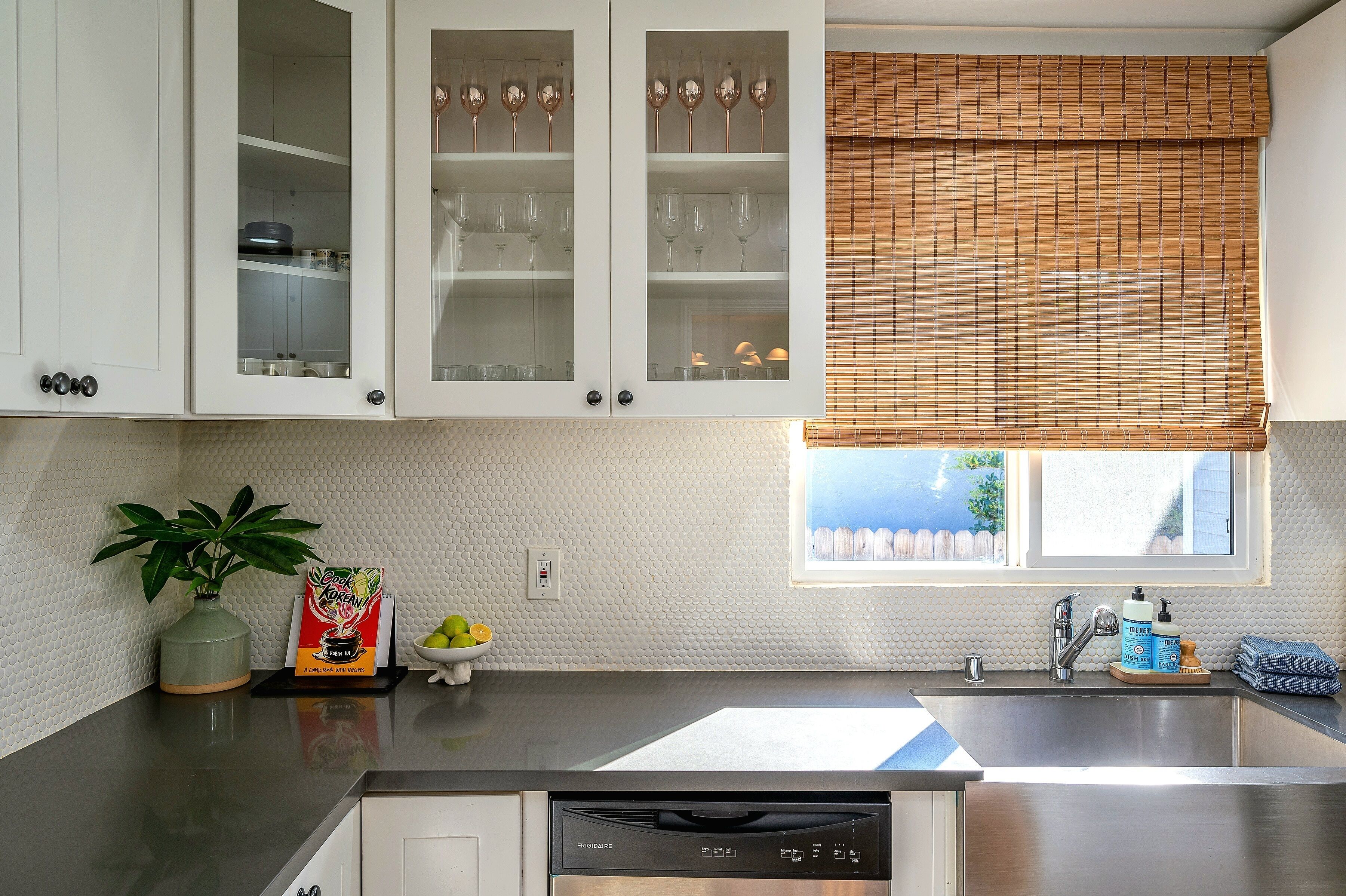 Una cocina con armarios blancos, un lavavajillas de acero inoxidable, una ventana con persiana de bambú y una planta en maceta sobre la encimera.