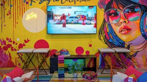 A vibrant room with a large TV displaying a scene, a neon sign, colorful chairs, a small table with a plant, and a colorful wall with a mural.