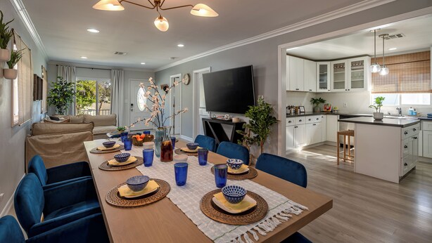 A modern dining area with a long table set for six, surrounded by blue chairs. A chandelier hangs above, and the room features a large TV and a kitchen area visible through an open doorway.