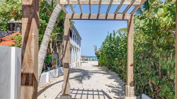 A beach pathway with a wooden pergola, surrounded by lush greenery and a clear view of the ocean.