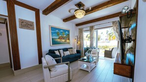 A living room with a dark blue sofa, a white armchair, a glass coffee table, wooden beams on the ceiling, a chandelier, and a painting of a beach scene.