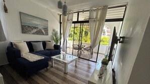 Un salon avec un canapé bleu foncé, une table basse blanche et un tableau accroché au mur. On y trouve une grande fenêtre avec des rideaux, une plante en pot et une petite table sur laquelle est posé un modèle de bateau.