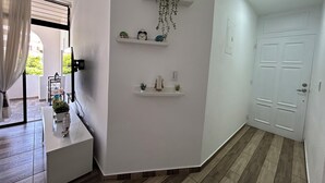 Un couloir aux murs blancs, doté d’un plancher en bois et d’une porte blanche. Des étagères avec des plantes et une horloge sont fixées au mur.
