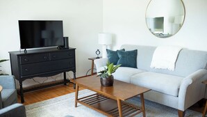 A modern living room with a flat-screen TV on a black TV stand, a light grey sofa with teal cushions, a wooden coffee table with a plant, a white lamp, and a round mirror on the wall. - The West Quincy House: In Town with Sauna + Garage (Hancock)