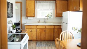 A compact kitchen with wooden cabinets, a white refrigerator, and a window with a white valance. - The West Quincy House: In Town with Sauna + Garage (Hancock)