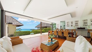 Uma sala de estar moderna com vista para o oceano, móveis de vime e um espaço de jantar com mesa de vidro.