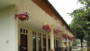 A building with a covered porch, hanging flower baskets, and a green barrel. - INDRA DJAYA HOTEL (Megamendung)