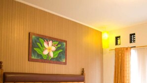 A neatly made bed with a headboard, a painting of a flower on the wall, and a window with curtains. - INDRA DJAYA HOTEL (Megamendung)