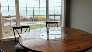 Una mesa redonda de madera con superficie de vidrio, rodeada por cuatro sillas, en una habitación con ventanas amplias con vista a una playa.