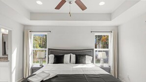 Une chambre avec un grand lit, deux tables de chevet avec des lampes, un ventilateur de plafond et des fenêtres avec des rideaux.