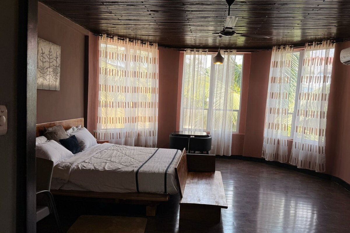Un dormitorio con cama, mesitas de noche, un banco y ventanas amplias con cortinas translúcidas.