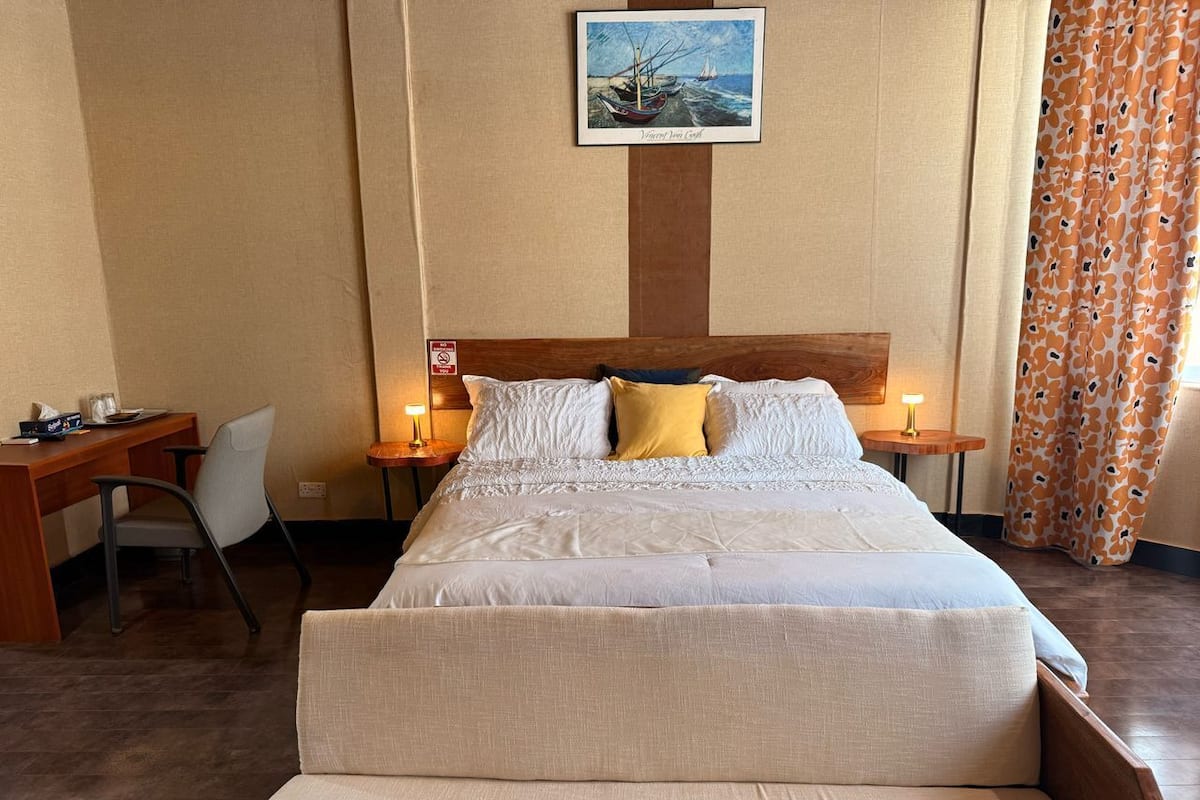 Ein Hotelzimmer mit einem großen Bett, zwei Nachttischen, einem Schreibtisch und einem Sessel. An der Wand hängt ein Gemälde, und das Vorhang hat ein Blattmuster.