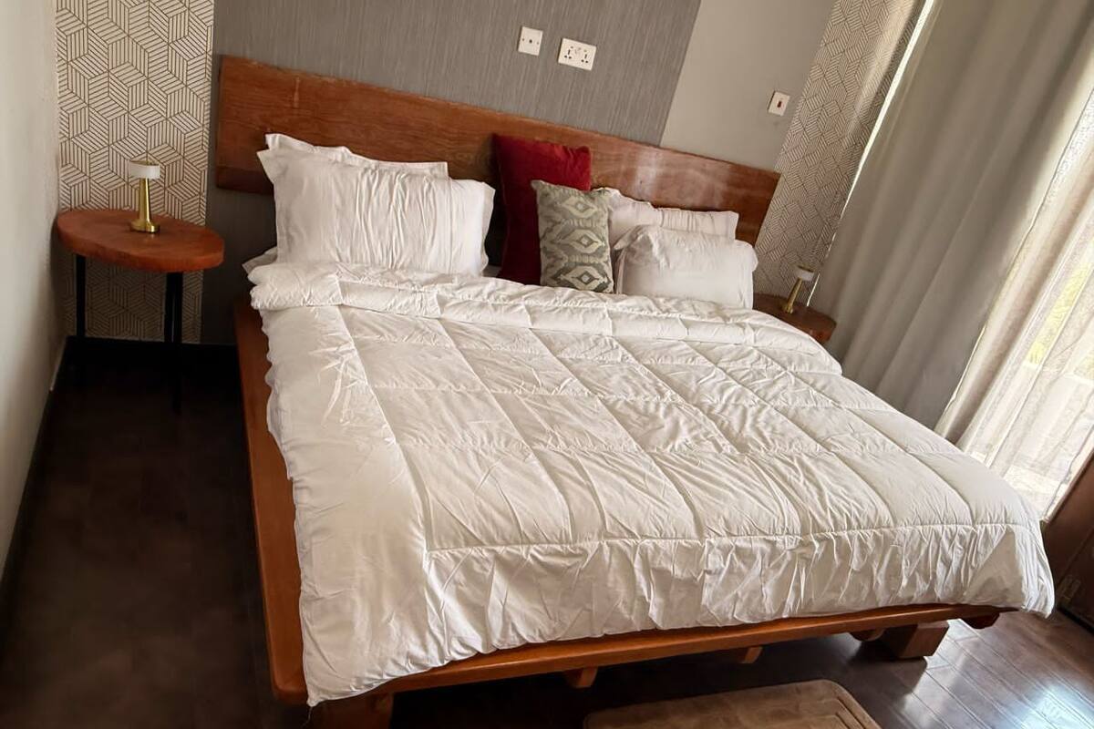 Ein großes Bett mit Holz Kopfteil, weißer Bettwäsche und mehreren Kissen. Neben dem Bett steht ein kleiner runder Tisch mit einer Lampe.