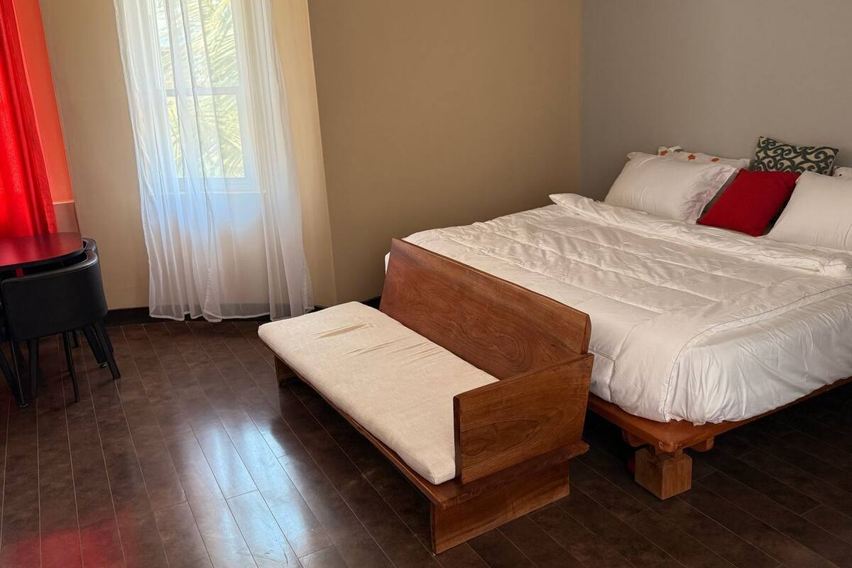 Ein Schlafzimmer mit einem hölzernen Bett, einer Bank, einem kleinen Tisch und einem Fenster mit durchsichtigen Vorhängen.