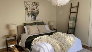 A neatly arranged bedroom with a bed, two bedside tables, a hanging light fixture, a painting, and a ladder against the wall.
