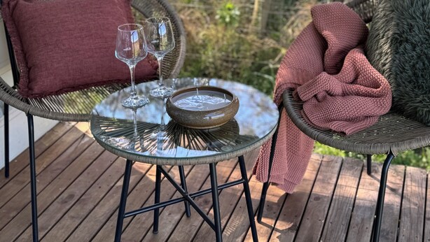 A wooden deck with two wicker chairs, one with a red cushion and the other with a green cushion. A glass table with a candle and a wine glass is in the center.