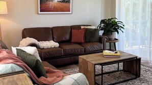 A living room with a brown leather sofa, a coffee table, and a large window with sheer curtains.