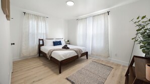 Un dormitorio con cama de madera, cortinas blancas, una mesita de noche pequeña y una planta.