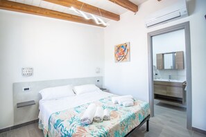 Una habitación de hotel moderna con cama, mesita de noche, aire acondicionado de pared, baño con espejo y lavamanos, y una obra de arte colorida en la pared.