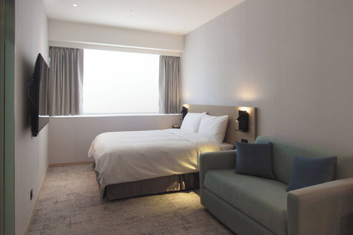 A hotel room with a bed, a green sofa, a TV mounted on the wall, and a window with curtains.
