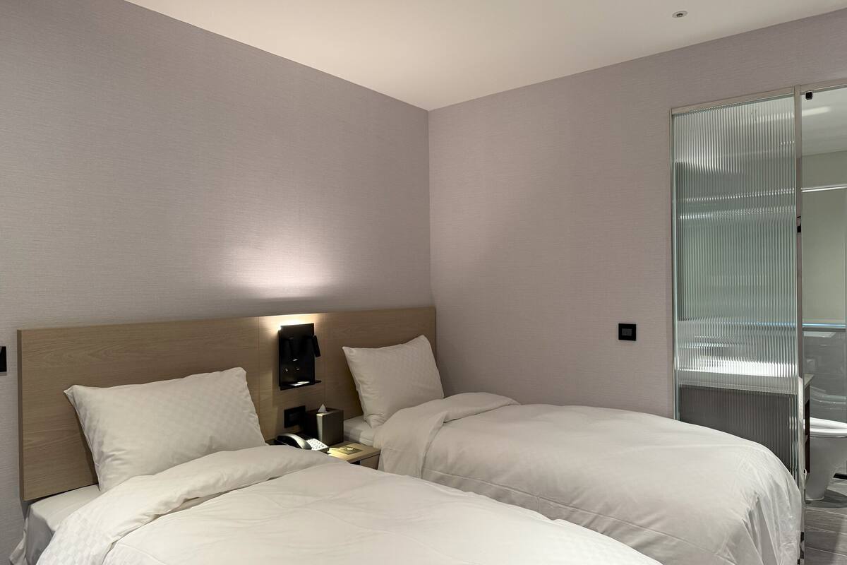 A hotel room with two beds, a bedside table with a lamp, and a sliding glass door leading to a bathroom.
