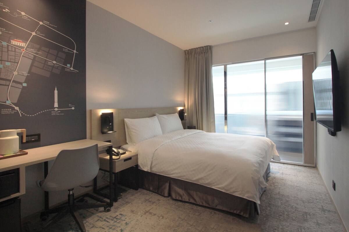 A hotel room with a bed, desk, chair, and a large window.