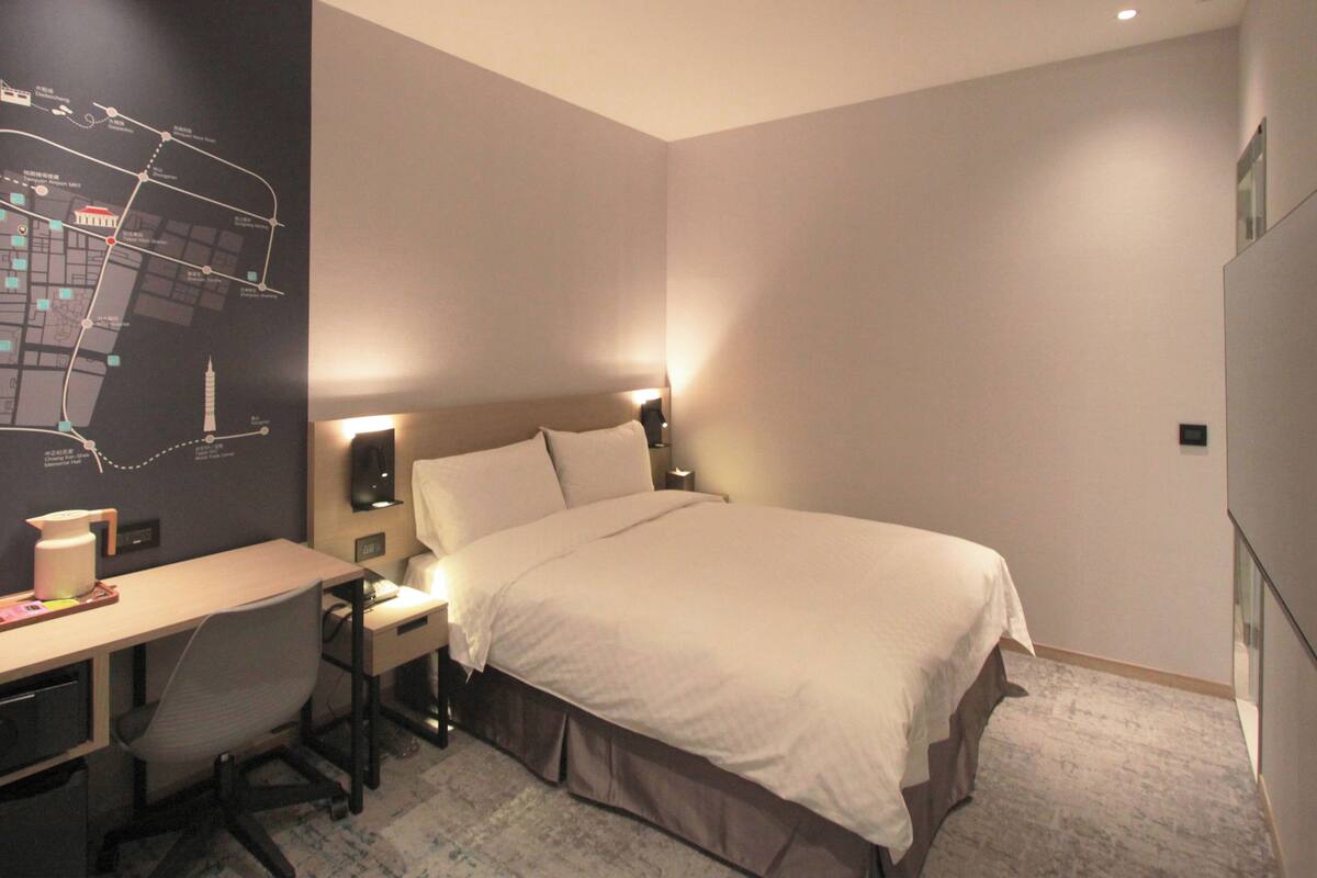 A hotel room with a bed, desk, chair, and a map on the wall.