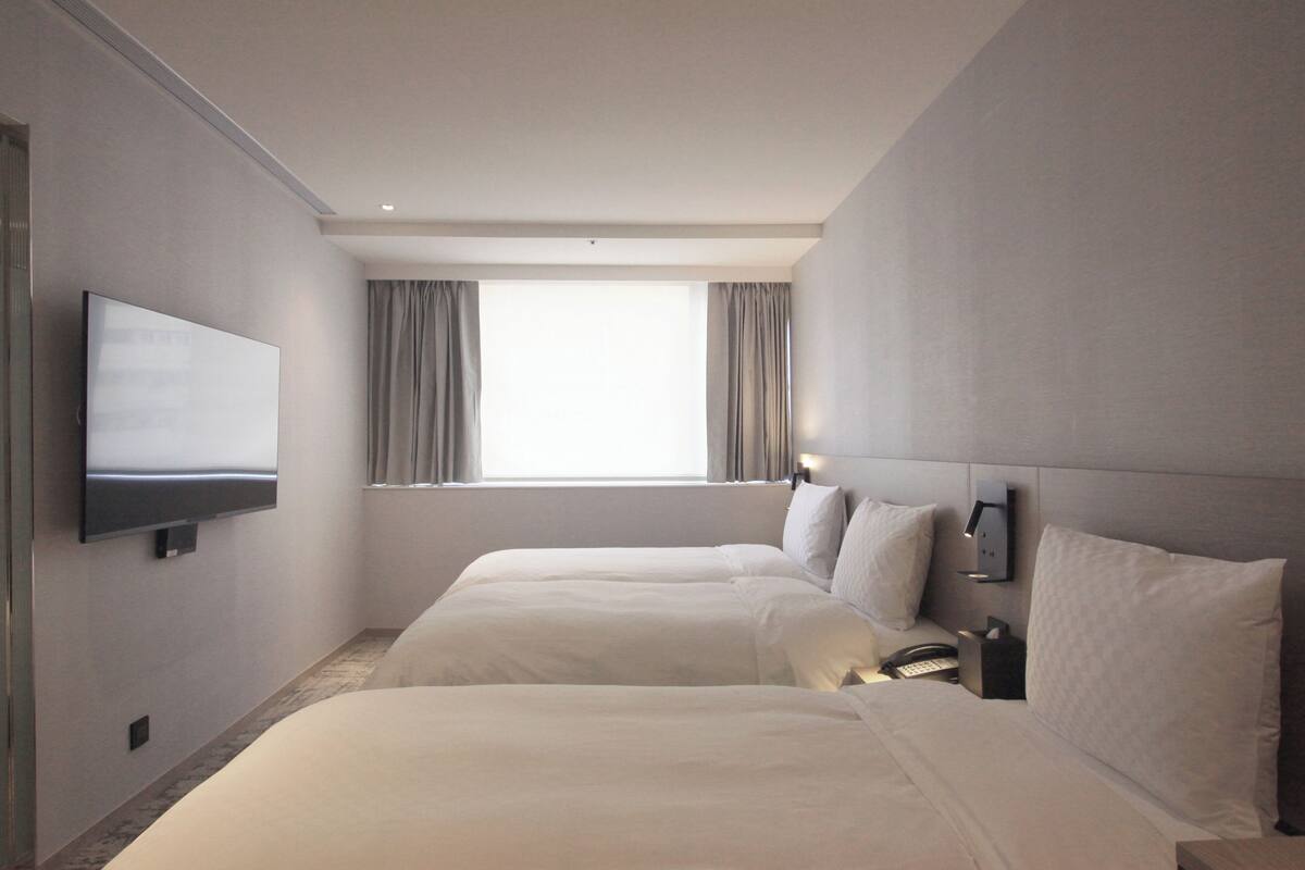 A hotel room with two beds, a flat-screen TV mounted on the wall, a bedside table with a lamp, and a window with curtains.