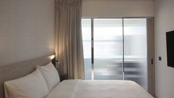 Una habitación de hotel con una cama grande, una puerta corrediza de vidrio a un balcón, un televisor de pantalla plana y una mesita de noche con lámpara.