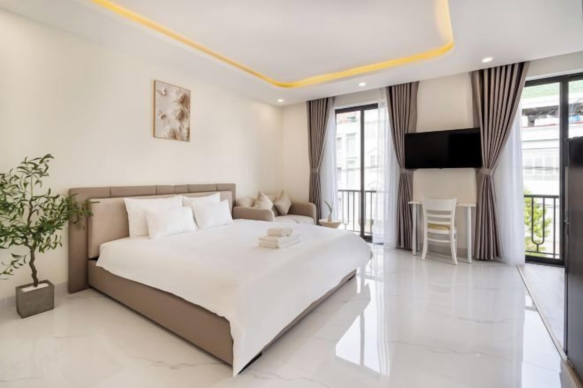 Una habitación de hotel moderna con una cama grande, una mesita de noche, una silla, un televisor montado en la pared y un balcón con cortinas.