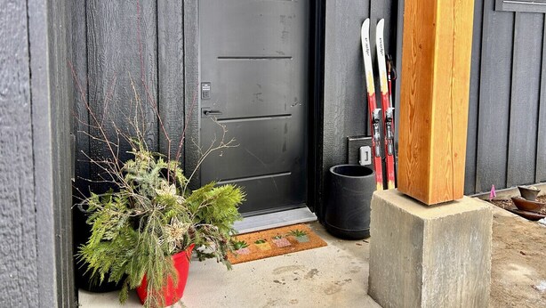 A black door with a small doormat, a potted plant, and skis leaning against the wall.