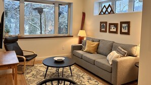 A living room with a sofa, a chair, a small table, and a lamp. There are windows with a view of trees and a lamp on the table.
