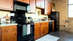 A kitchen with wooden cabinets, a black stove, a refrigerator, and a brick wall. - The Luxe Loft in Canal Park | Boho-Chic (Duluth)