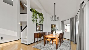 A modern dining area with a wooden table and chairs, a chandelier, and a view of the outdoors through large windows.