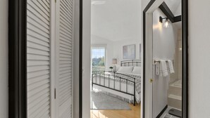 A bedroom with a bed, a nightstand, a window, and a bathroom with a shower and a towel rack.