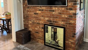 A living room with a brick fireplace, a flat-screen TV mounted above it, and a framed picture below. -  NO CLEANING FEE Century farmhouse on Lake Keowee near Clemson (Seneca)