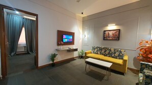 A modern living room with a yellow sofa, a small white table, a mounted TV, and a painting on the wall. - Mallenzo suites (Gurugram)