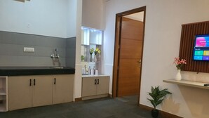 A modern kitchen with a black countertop, a sink, and cabinets. There is a wooden door leading to another room. A white table with a glass bowl is in the foreground. - Mallenzo suites (Gurugram)
