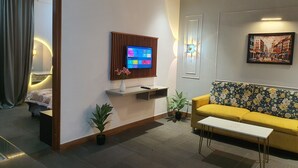 A modern living room with a flat-screen TV mounted on a wooden panel, a sofa, a small table, and a potted plant. - Mallenzo suites (Gurugram)