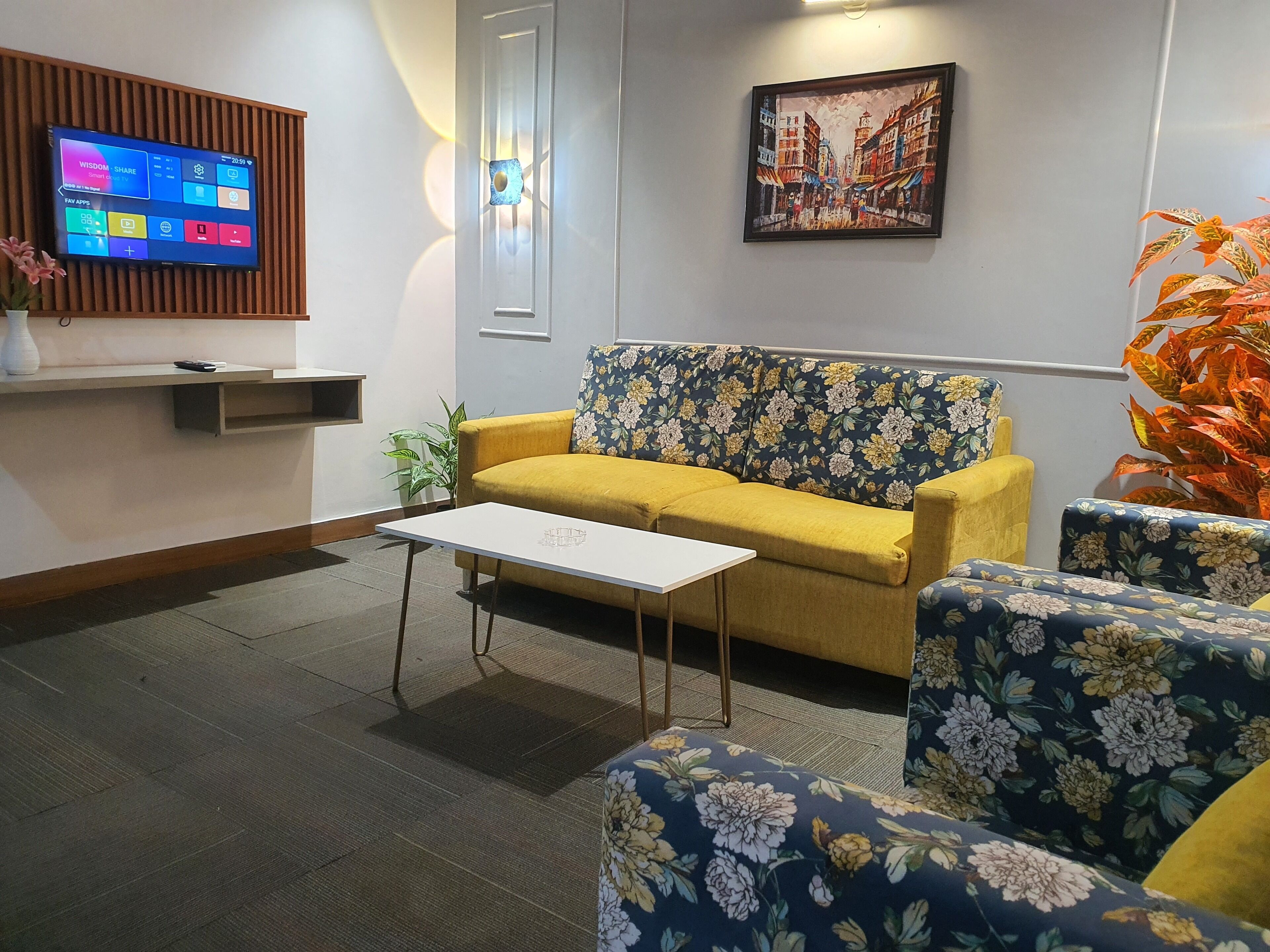 A modern living room with a yellow sofa, floral-patterned armchairs, a flat-screen TV mounted on a wooden panel, a framed cityscape painting, and a small table.