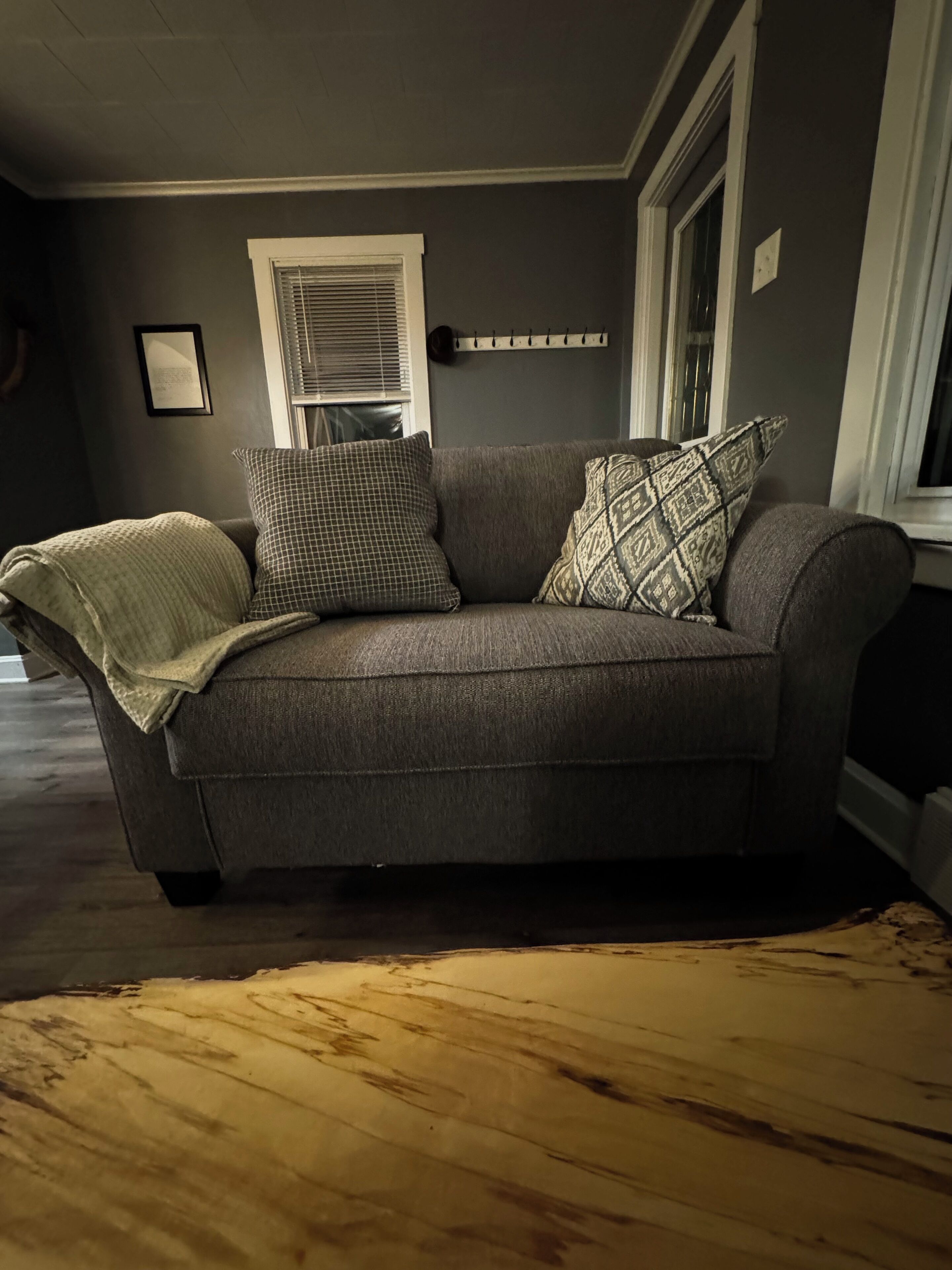 Un sofá gris con cojines estampados y una manta, ubicado en una habitación con piso de madera y una ventana con persianas.