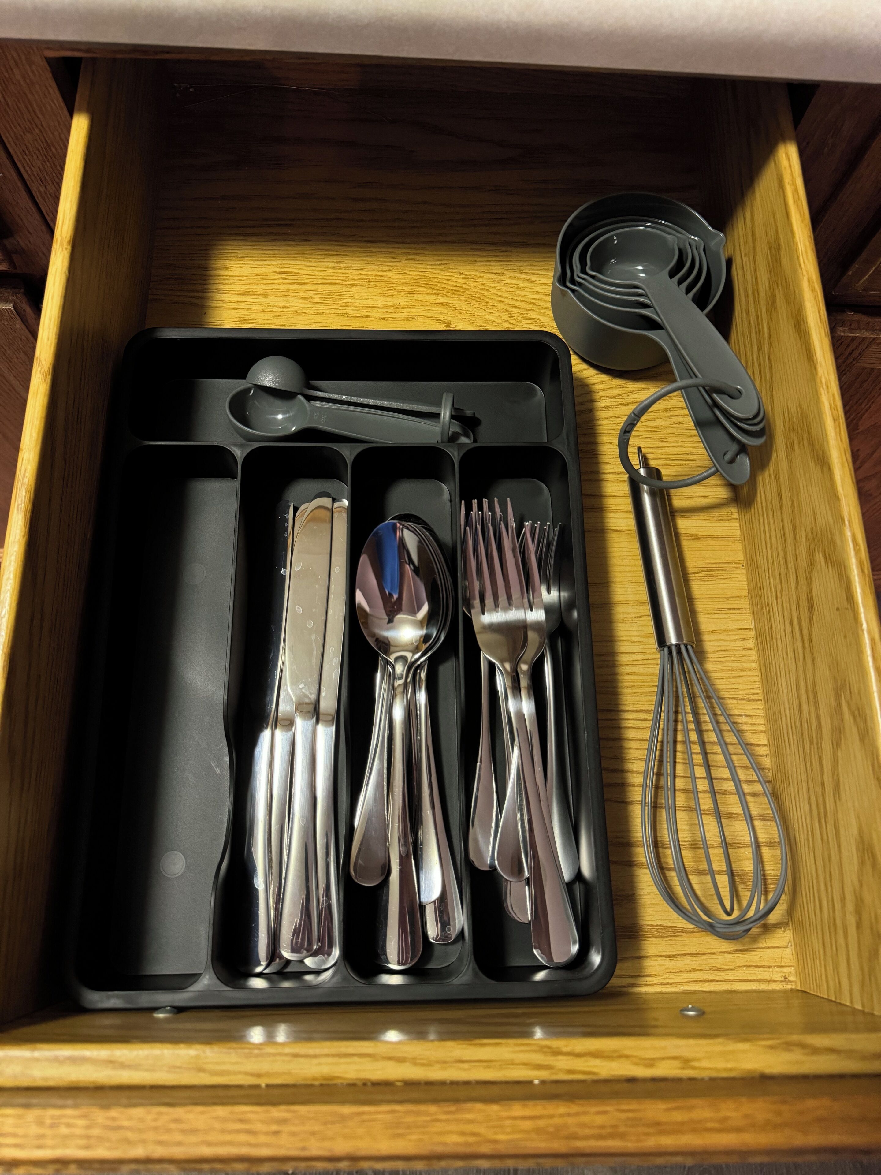 A drawer containing a black tray with silverware, a whisk, and a measuring cup.