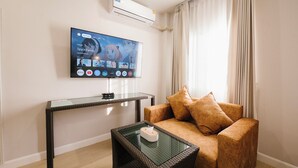 A living room with a flat-screen TV mounted on the wall, a glass coffee table, a brown sofa, and a wicker side table. - Eng Fah Luang Hotel (Chiang Rai)
