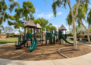 A playground with slides and climbing structures, surrounded by trees and a clear sky.
