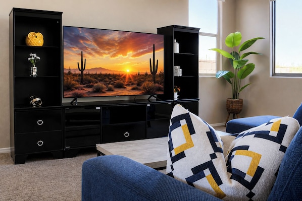 A living room with a flat-screen TV, black entertainment unit, blue sofa, patterned cushions, and a potted plant.