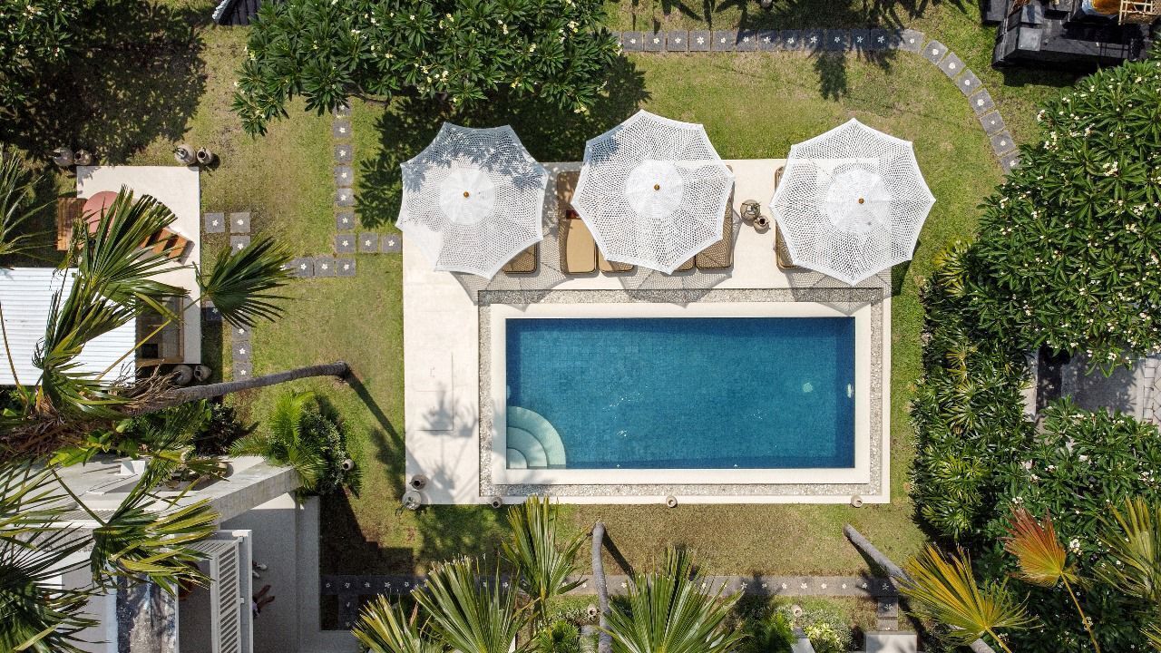An aerial view of a swimming pool with three white umbrellas, surrounded by well-maintained greenery and a paved walkway.