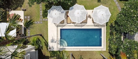 Vista aérea de una piscina con tres sombrillas blancas, rodeada de vegetación cuidada y un paseo empedrado.